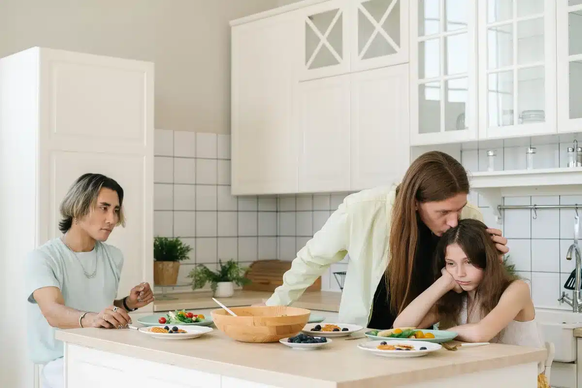 Padre preocupado sentado en la cocina con papeles y café, luz natural de ventana iluminando su expresión pensativa.