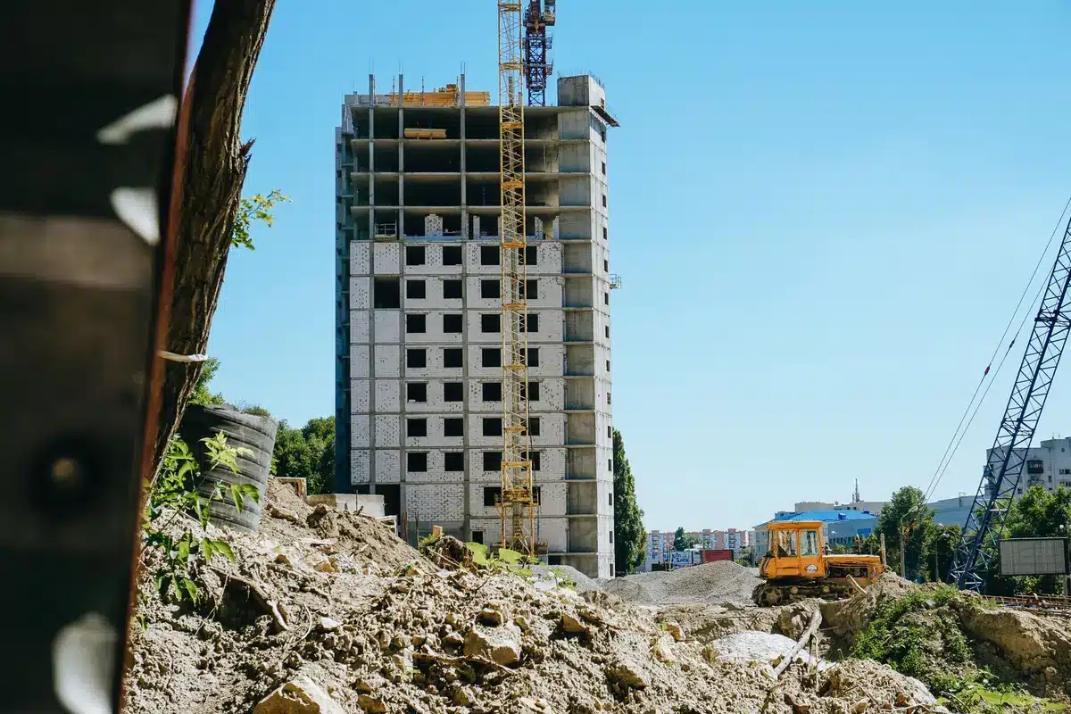 Anonymous construction site with mid-rise scaffolding and a crane beside completed low-rise housing, empty street in foreground suggesting potential redevelopment plots