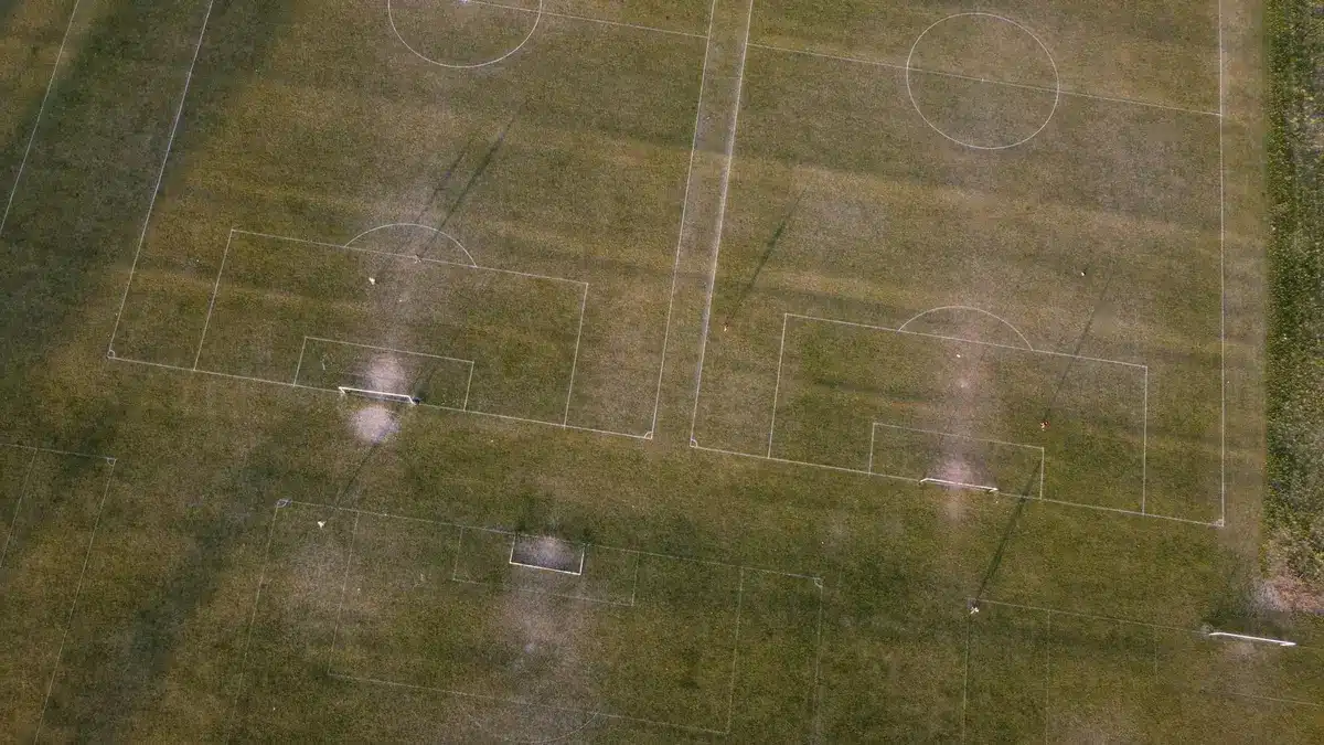 A distant view of an empty football pitch at dusk, a lone goal and scattered training cones, stadium lights on in the background, mood of quiet preparation