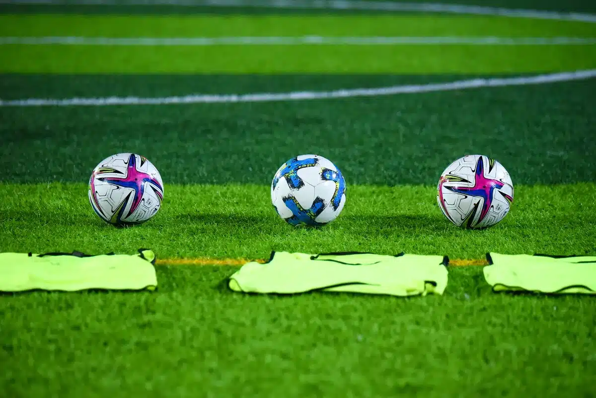 Training bibs and a plain white ball lying on worn turf, an anonymous pair of gloves beside the ball suggesting recent action, empty stands blurred beyond the touchline