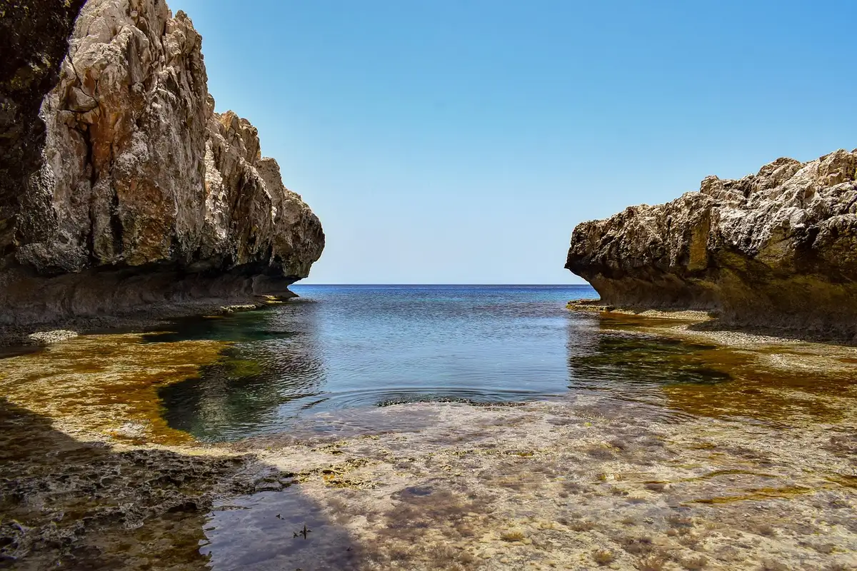 A deserted rocky cove at golden hour with clear turquoise water lapping a small sandy inlet, weathered cliffs framing the scene, no people or signage