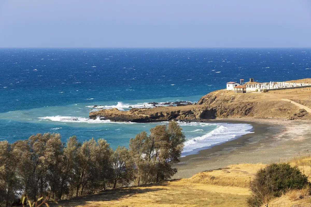 Aerial view of a narrow sandy beach backed by scrubland and low cliffs, a single narrow path leading down from a dirt track, empty but clearly accessible