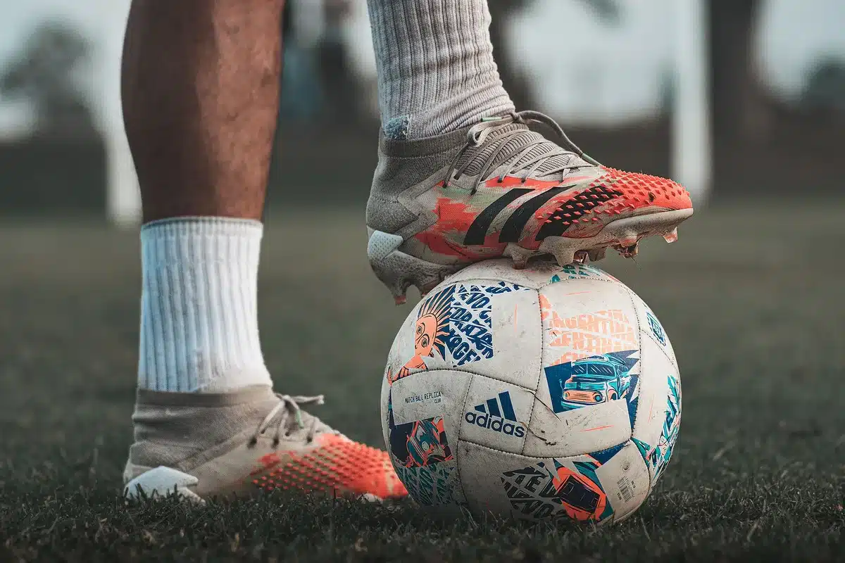 Close-up of an unbranded football boot on the sideline beside a match ball, grass blades and pitch paint visible