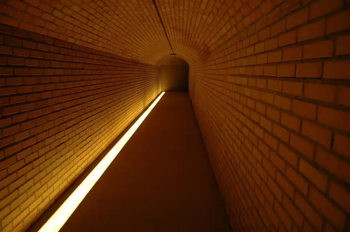 An empty vaulted brick passage lit by warm lamps, a straight arched corridor receding into shadow with scattered historic-style iron lanterns and damp stone floor