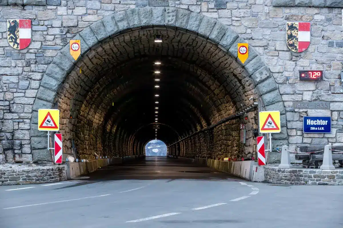 Wide shot of an unbranded stone tunnel entrance lit from inside; no people, damp floor, moss on the walls, a small plaque holder empty beside the arch