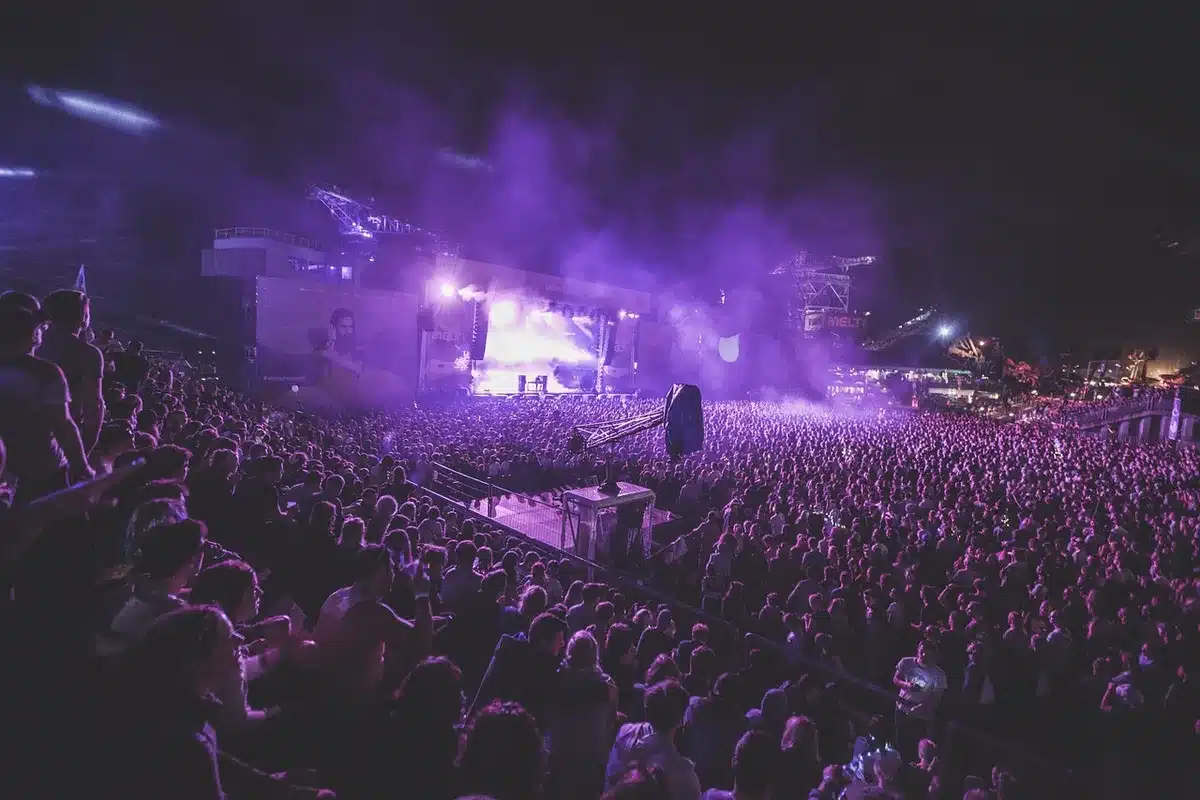 Wide shot of an indoor arena stage after a concert; empty instruments on stage, scattered confetti on the floor, distant anonymous crew moving under warm lights