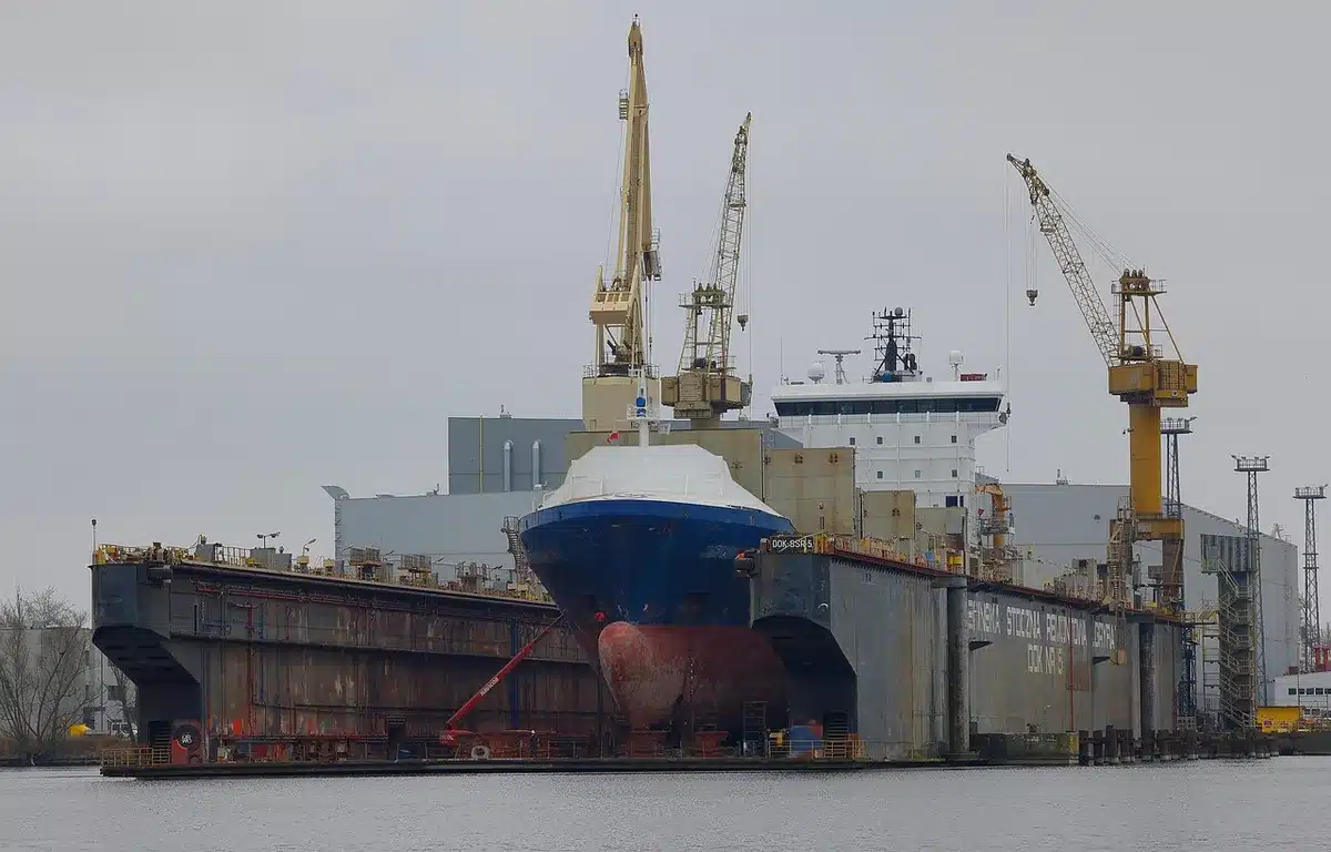 Wide view of an industrial dock with cranes and an anonymous crowd area for a ceremony, overcast sky but clear details