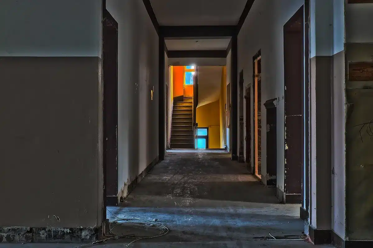 An empty, decayed concrete corridor with scattered debris and a rusted gas mask on the floor, light streaming through broken windows