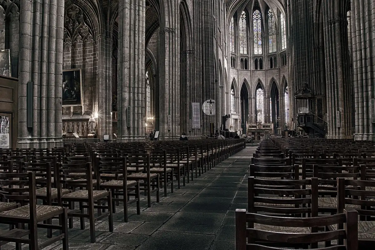 An empty cathedral nave with wooden pews, scattered floral arrangements along the aisle, soft daylight through stained-glass windows, an unbranded horse bridle on a side bench
