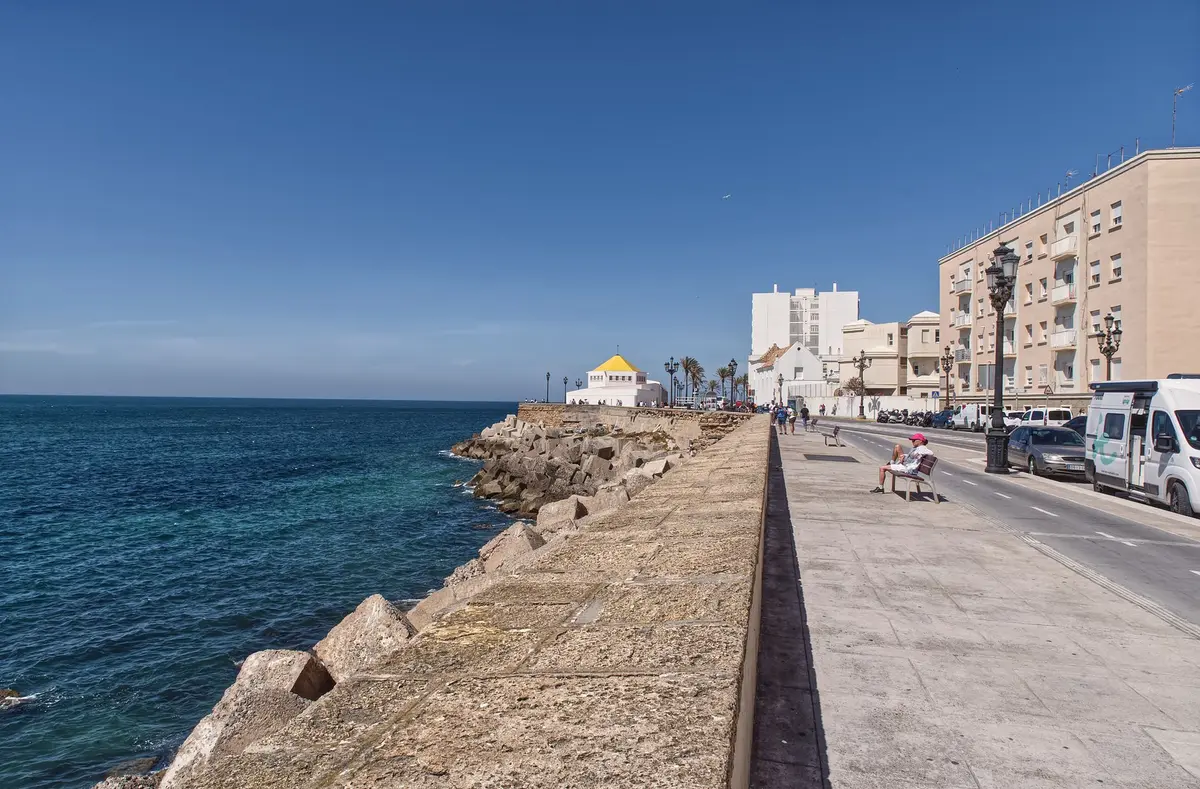 Empty coastal promenade at dusk with a lone bench and distant low buildings, no people
